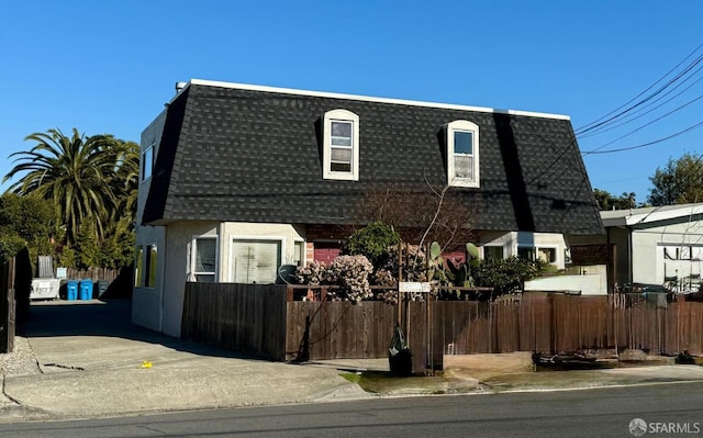 view of property with mansard roof, a shingled roof, fence, and stucco siding