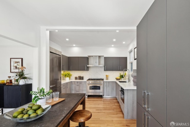 kitchen with light wood finished floors, a sink, stainless steel appliances, wall chimney range hood, and modern cabinets