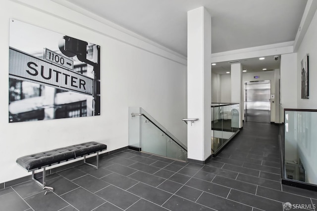 corridor featuring elevator, baseboards, dark tile patterned flooring, and an upstairs landing