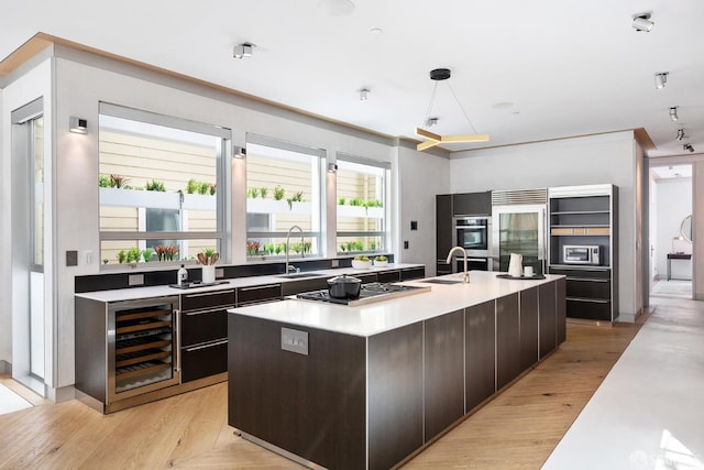 kitchen with wine cooler, an island with sink, appliances with stainless steel finishes, light hardwood / wood-style floors, and decorative light fixtures