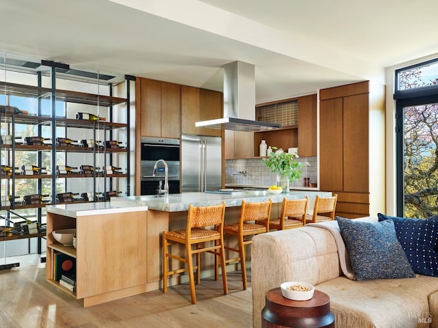 kitchen featuring stainless steel built in refrigerator, tasteful backsplash, island range hood, light wood-type flooring, and black double oven