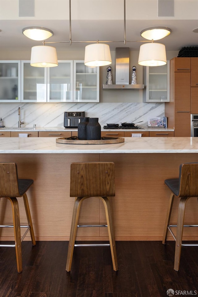 kitchen featuring wall chimney exhaust hood, a kitchen bar, glass insert cabinets, and dark wood-type flooring