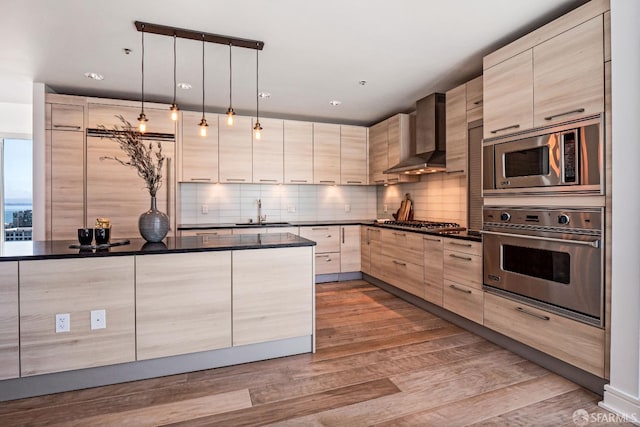 kitchen with light brown cabinets, decorative light fixtures, wall chimney exhaust hood, appliances with stainless steel finishes, and light hardwood / wood-style floors