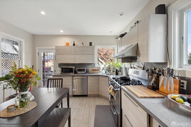 kitchen with decorative backsplash, modern cabinets, appliances with stainless steel finishes, light brown cabinetry, and under cabinet range hood
