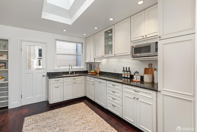 kitchen with a skylight, a sink, dark stone counters, stainless steel microwave, and dark wood finished floors