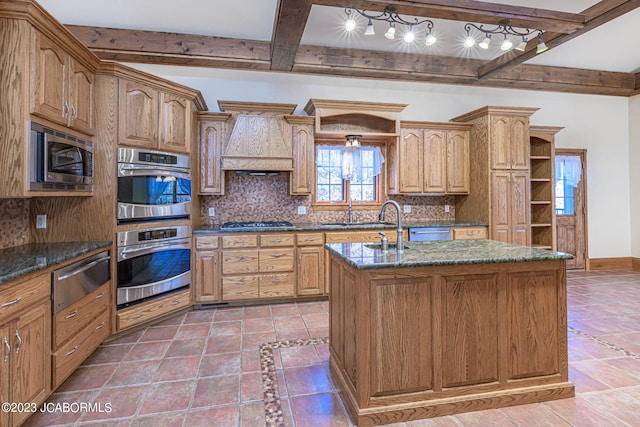 kitchen featuring tasteful backsplash, premium range hood, stainless steel appliances, beam ceiling, and a center island with sink