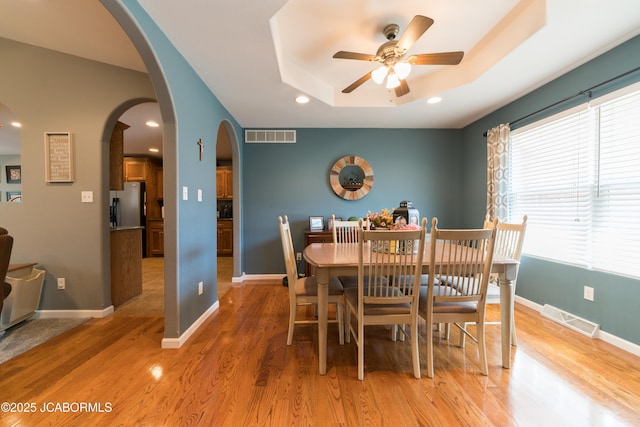 dining room featuring visible vents, light wood-style floors, arched walkways, a raised ceiling, and a ceiling fan