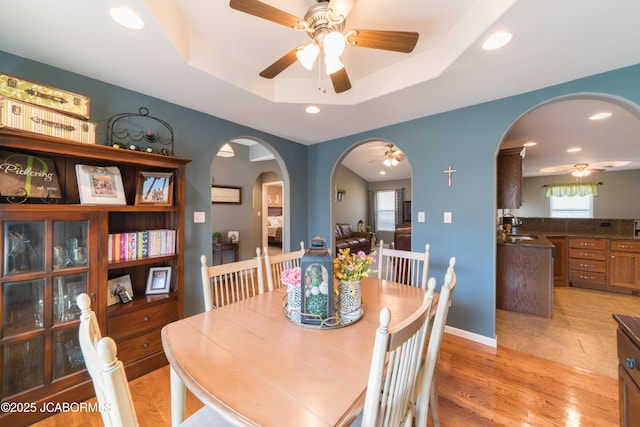 dining space with a tray ceiling, light wood-style floors, arched walkways, and ceiling fan