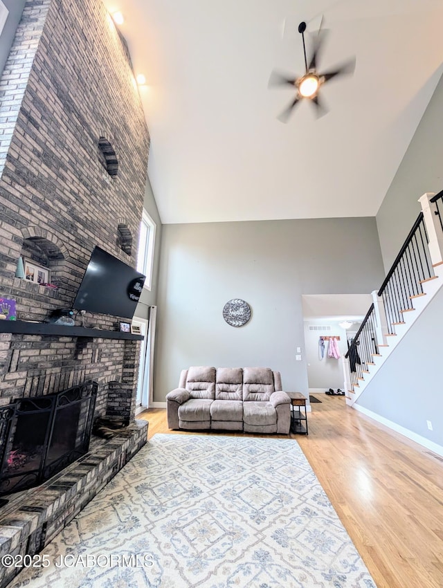 living area featuring a fireplace, wood finished floors, a towering ceiling, a ceiling fan, and stairs