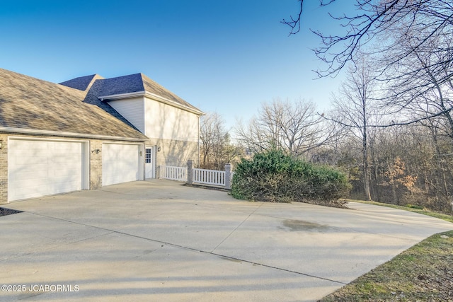 view of side of property featuring driveway, fence, roof with shingles, a garage, and brick siding