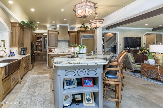 kitchen featuring a spacious island, wall chimney exhaust hood, decorative backsplash, light stone counters, and a kitchen bar