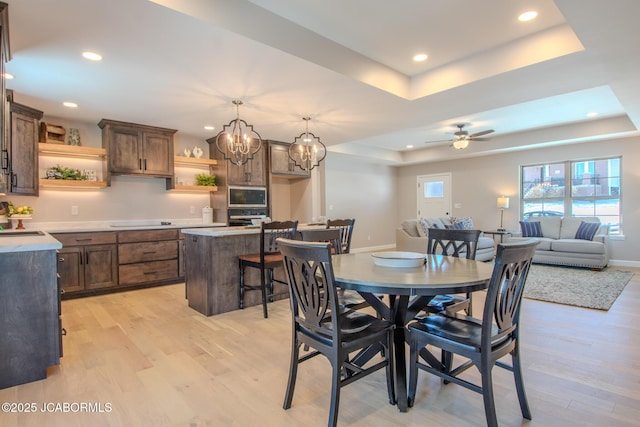 dining room with a raised ceiling, sink, ceiling fan with notable chandelier, and light hardwood / wood-style flooring