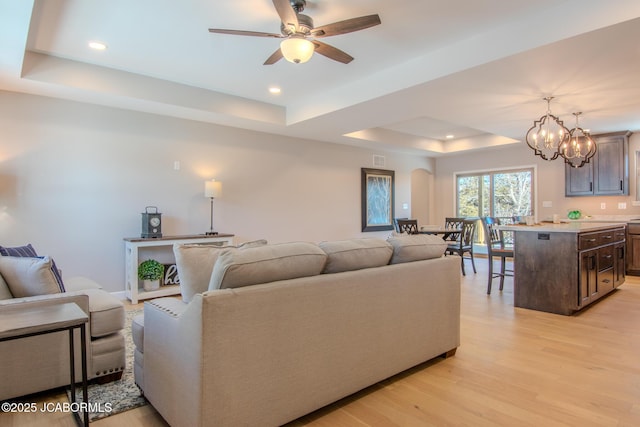 living room featuring a raised ceiling, ceiling fan with notable chandelier, and light hardwood / wood-style flooring