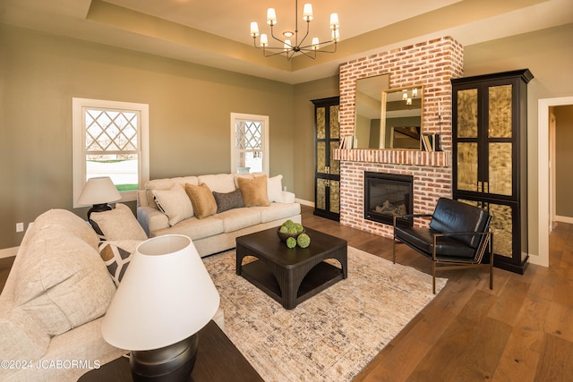 living room featuring a raised ceiling, wood-type flooring, a notable chandelier, and a brick fireplace