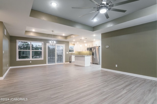 unfurnished living room featuring a raised ceiling, ceiling fan with notable chandelier, and light wood-type flooring