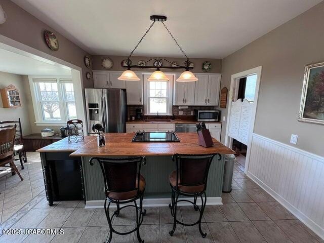 kitchen featuring stainless steel appliances, light tile patterned floors, wainscoting, white cabinetry, and decorative light fixtures