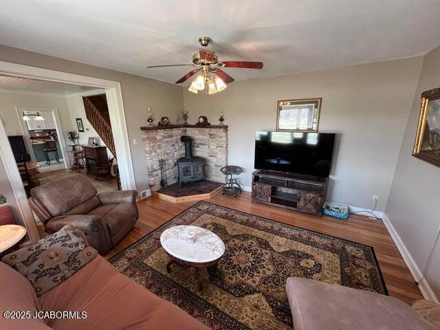living room featuring wood finished floors, a wood stove, stairway, a ceiling fan, and baseboards