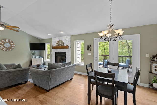 dining room featuring lofted ceiling, a stone fireplace, baseboards, light wood finished floors, and ceiling fan with notable chandelier