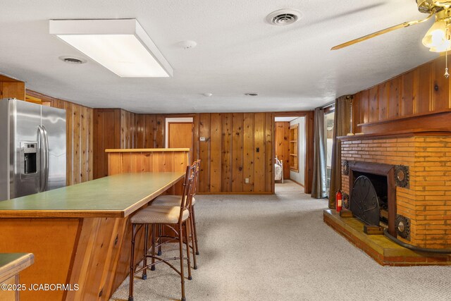 24671 Gardner Rd, Versailles, MO 65084 - kitchen with stainless steel refrigerator with ice dispenser, brown cabinetry, wood walls, carpet, and a brick fireplace