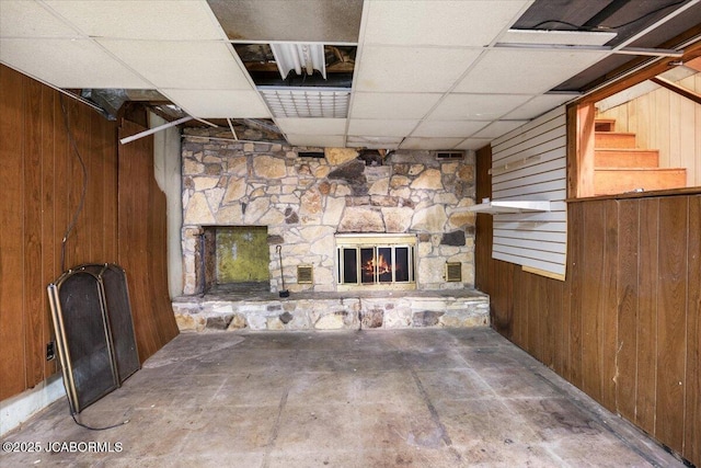 unfurnished living room with a drop ceiling, a stone fireplace, and wooden walls