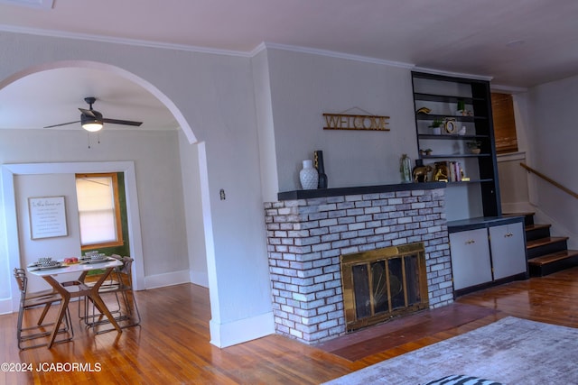 living room featuring crown molding, built in shelves, ceiling fan, a fireplace, and wood-type flooring