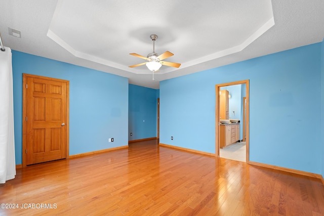 unfurnished bedroom featuring ensuite bathroom, light hardwood / wood-style flooring, ceiling fan, a textured ceiling, and a tray ceiling