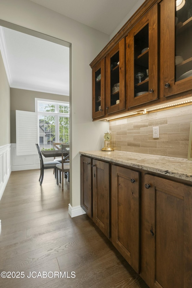 kitchen with tasteful backsplash, light stone countertops, glass insert cabinets, wainscoting, and dark wood-type flooring