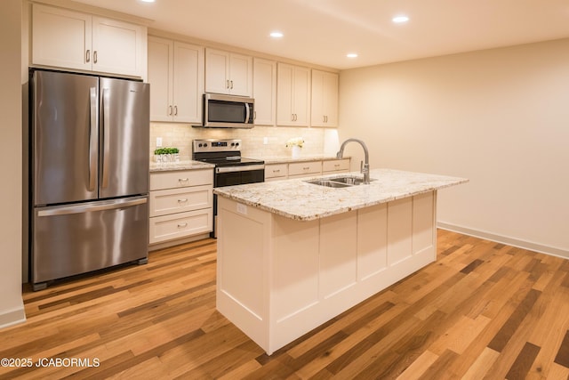 kitchen featuring a kitchen island with sink, stainless steel appliances, a sink, light wood-type flooring, and light stone countertops