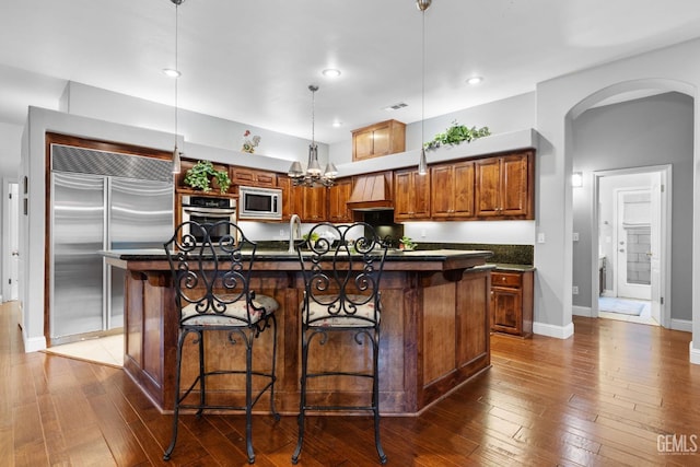 kitchen featuring pendant lighting, dark wood-type flooring, built in appliances, an island with sink, and a kitchen bar