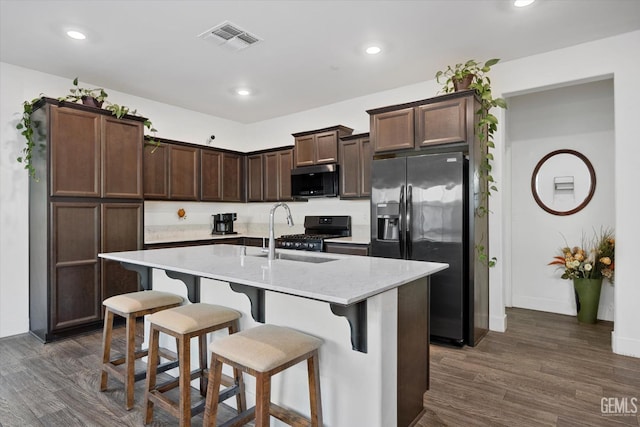 kitchen featuring an island with sink, sink, stainless steel appliances, light stone countertops, and dark wood-type flooring