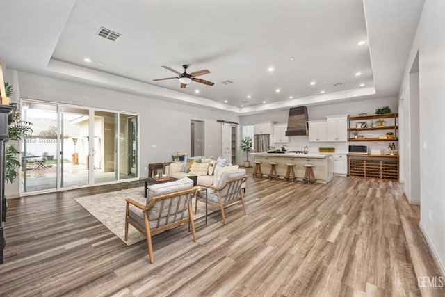 living room with light wood-type flooring, a tray ceiling, visible vents, and ceiling fan