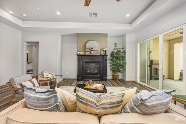 living area featuring visible vents, wood finished floors, a glass covered fireplace, a raised ceiling, and a ceiling fan