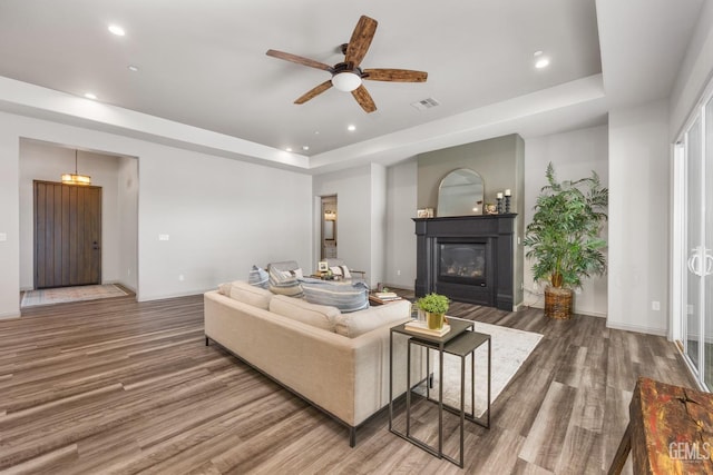 living room featuring visible vents, a tray ceiling, wood finished floors, a glass covered fireplace, and a ceiling fan