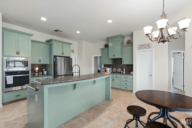 kitchen featuring visible vents, hanging light fixtures, appliances with stainless steel finishes, an island with sink, and dark stone counters