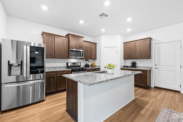 kitchen featuring dark brown cabinets, appliances with stainless steel finishes, light stone countertops, a center island with sink, and light hardwood / wood-style floors