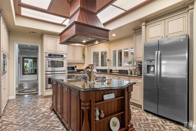 kitchen featuring appliances with stainless steel finishes, brick floor, cream cabinetry, visible vents, and custom exhaust hood