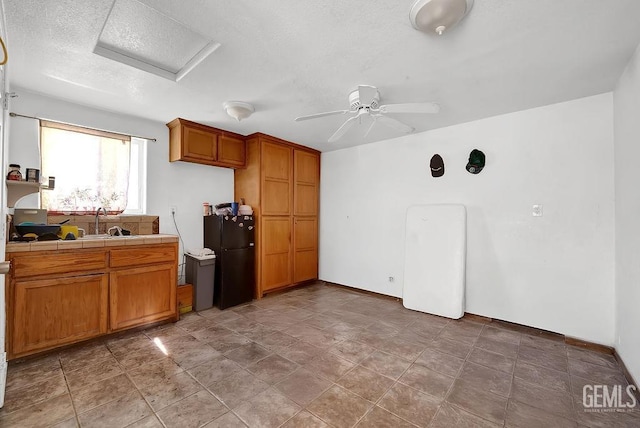 kitchen with tile counters, brown cabinets, a sink, freestanding refrigerator, and a textured ceiling