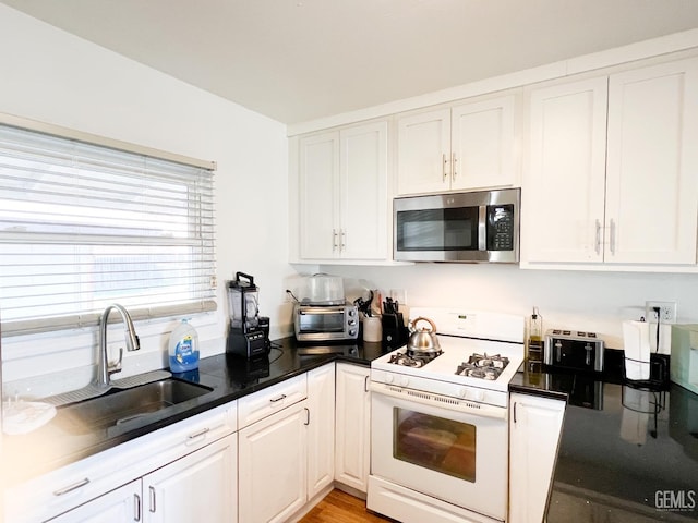 kitchen featuring white range with gas cooktop, a sink, stainless steel microwave, dark countertops, and white cabinets