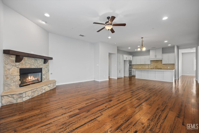 unfurnished living room with ceiling fan, a stone fireplace, visible vents, baseboards, and dark wood-style floors