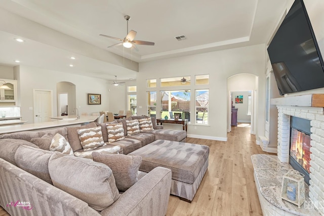 living room with a brick fireplace, light hardwood / wood-style flooring, a raised ceiling, and ceiling fan