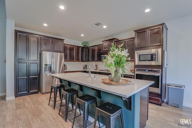 kitchen with a center island with sink, appliances with stainless steel finishes, light hardwood / wood-style floors, a kitchen bar, and dark brown cabinetry