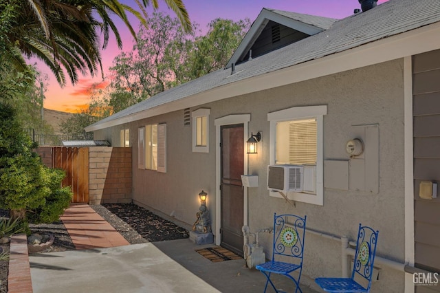 exterior entry at dusk with a shingled roof, cooling unit, fence, and stucco siding