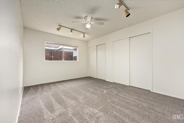 unfurnished bedroom featuring ceiling fan, rail lighting, a textured ceiling, carpet flooring, and two closets