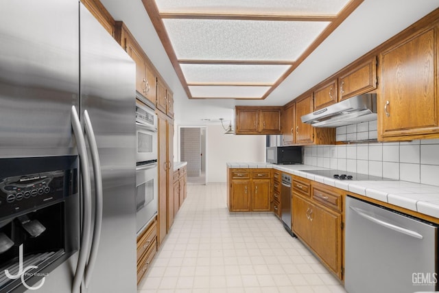 kitchen with brown cabinetry, tile countertops, tasteful backsplash, black appliances, and under cabinet range hood