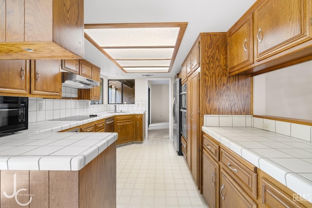 kitchen featuring decorative backsplash, tile counters, under cabinet range hood, a sink, and black appliances