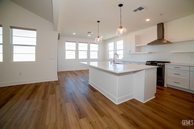 kitchen featuring stainless steel gas stove, white cabinetry, hardwood / wood-style flooring, a center island with sink, and wall chimney exhaust hood