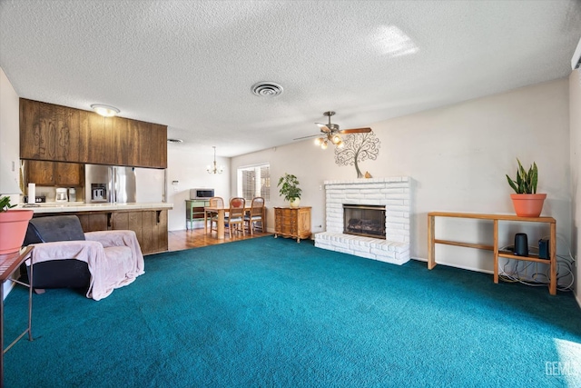 carpeted living room featuring ceiling fan with notable chandelier, a brick fireplace, and a textured ceiling