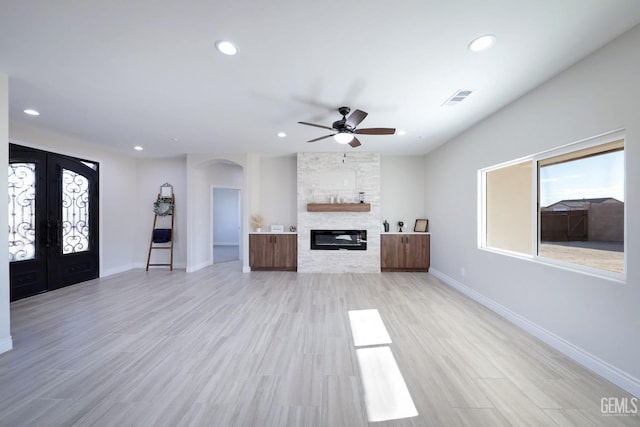 unfurnished living room with a stone fireplace, ceiling fan, french doors, and light wood-type flooring