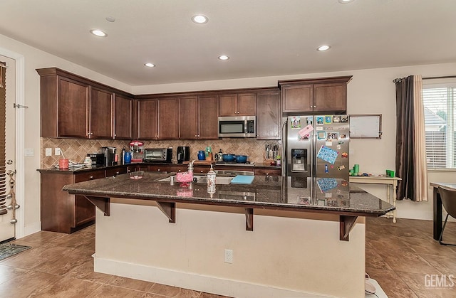 kitchen with stainless steel appliances, a breakfast bar, dark stone countertops, and a center island with sink