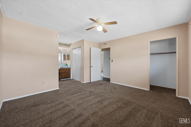 unfurnished bedroom featuring ensuite bathroom, dark carpet, a textured ceiling, ceiling fan, and multiple closets